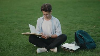 Genç adam kitap okuyor ve dışarıda notlar alıyor. Üniversite öğrencisi parkta okuyor. Eğitim, öğrenme, bilgi.