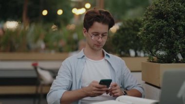 Genç bir adam dışarıda bir kafede çalışırken akıllı telefon kullanıyor. Telefonuna ve işine odaklanmış durumda..
