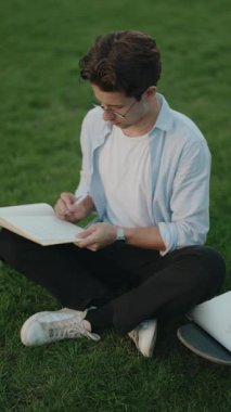 Genç adam parkta çimlere oturur ve deftere yazar. Gözlük, beyaz gömlek ve siyah pantolon giyiyor. Kaykayı arkasında..