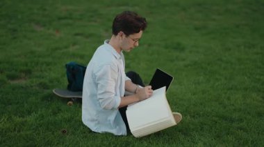 Genç bir adam çimenlerde oturur, çalışmalarını çalışır. Dizüstü bilgisayarına gönderme yaparken büyük bir deftere not alır..