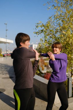 Açık havada iki insan, açık bir gökyüzü altında antrenman yapmaya hazırlanırken esneme hareketleri yapıyor..