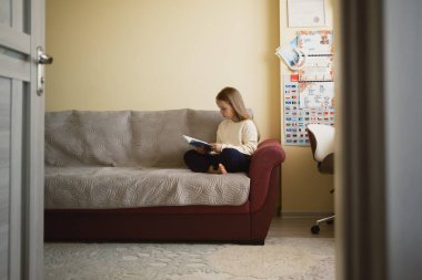 Kanepede oturup kitap okuyan bir kız. Çalışma odası olan rahat bir odada..