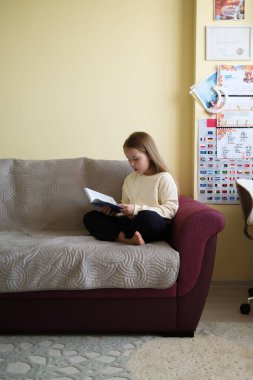 Genç bir kız kitap okurken bir kanepede oturuyor. Oda sıcak bir şekilde aydınlatılmış, öğrenme ve sakinleştirme atmosferi sergiliyor. Eğitimsel materyaller görünür olup, çalışkan bir arkaplan eklenir.