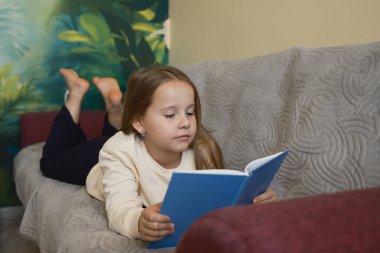 Genç bir kız rahat bir kanepede uzanıyor, dikkatle mavi bir kitap okuyor. Rahat ve sakin atmosfer, onun huzurlu bir çevredeki hareketliliğini vurguluyor..