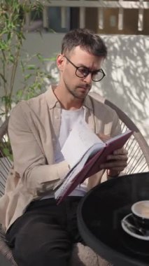 Genç bir adam balkonunda oturuyor, defterinde çalışıyor ve bir fincan kahvenin tadını çıkarıyor. O, gözlük, açık kahverengi bir gömlek ve koyu pantolon giyiyor..