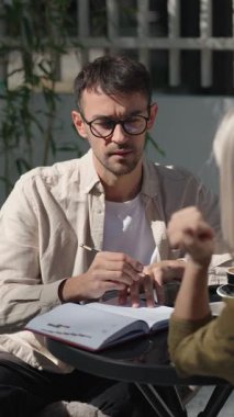 Bir adam ve bir kadın bir masada birlikte oturuyorlar ve bir kafede iş toplantısı yapıyorlar. Not defterine notlar yazıyor ve konuşuyor..