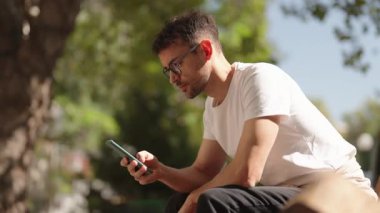Gözlüklü genç bir adam parkta bir bankta oturuyor ve telefonuna bakıyor. Beyaz tişört ve siyah pantolon giyiyor..