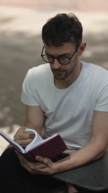 Şehir parkında kitap okuyan bir adam. Beyaz bir gömlek ve gözlük takıyor ve sessiz vakit geçiriyor..