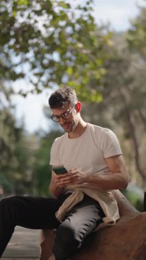 Genç bir adam parkta bir kütüğe oturmuş, telefonunu kullanırken gülümsüyor. Beyaz tişört, siyah pantolon, gözlük takıyor ve beline bej bir tişört bağlanmış..