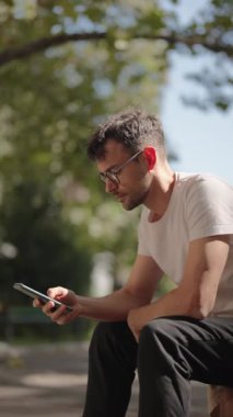 Genç bir adam güneşli bir yaz günü parkta bankta otururken akıllı telefonunu kontrol eder. Günlük kıyafetler ve gözlük takıyor..