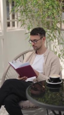 Adam kitap okuyor ve açık bir kafede kahve içiyor. Bej bir gömlek ve gözlük takıyor, rahat bir sandalyede oturuyor..