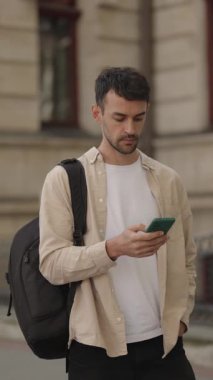 Bej bir tişört ve sırt çantası giyen genç bir adam telefonunu kontrol ediyor. Şehir caddesinde bir binanın önünde duruyor..