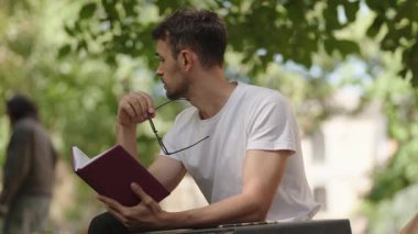 Düşünceli bir adam parkta bir bankta oturuyor, bir elinde gözlüğü, diğer elinde bir defter, sonraki hamlesini düşünüyor..