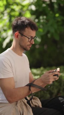 Genç bir adam parkta oturuyor, kamerasındaki fotoğrafları inceliyor. Sonuçlardan memnun görünüyor..