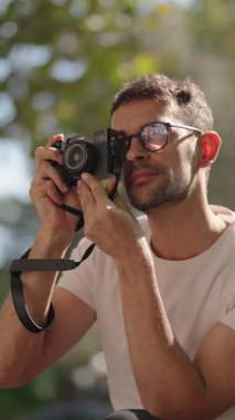 Gözlüklü ve beyaz tişörtlü genç bir adam klasik bir kamerayla dışarıda fotoğraf çekiyor. Fotoğrafçılık konusunda tutkuludur ve özel anları yakalamayı sever..