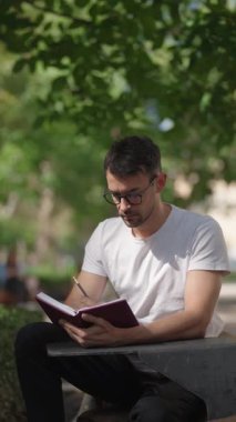 Adam dışarıda oturup deftere bir şeyler yazıyor. Gözlük, beyaz tişört ve siyah pantolon giyiyor. Dışarıda bir masada oturur ve ağacın gölgesinden zevk alır..