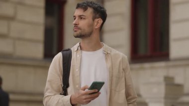 Genç bir adam telefonunu tutar ve etrafa bakar. O, dışarıda büyük eski bir binanın önünde duruyor. Beyaz bir tişört ve siyah bir sırt çantası giyiyor..