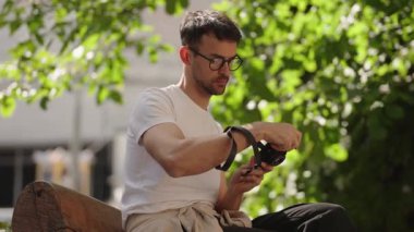 Genç bir adam parkta bir bankta oturuyor, kamerasını ayarlıyor. Gözlük, beyaz tişört ve siyah pantolon giyiyor. Güneşli bir gün ve yemyeşil yeşil huzurlu bir atmosfer yaratır..