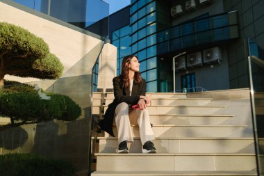 Yumuşak güneş ışığında modern bir mimariye karşı düşünceli adımlarla oturan bir kadın.