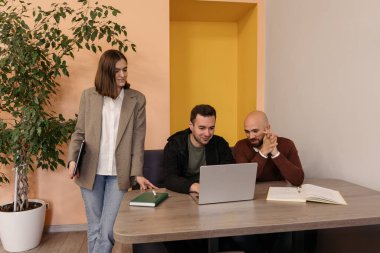 Modern ofis ortamındaki bir dizüstü bilgisayara odaklanmış üç iş arkadaşı iş hakkında konuşuyor.