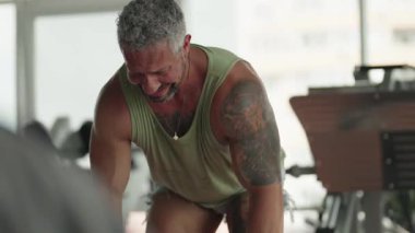 Dövmeleri olan kaslı bir adam spor salonunda dambıllarla egzersiz yapar, gücünü gösterir ve odaklanır..