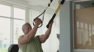 Modern bir spor salonunda süspansiyon kayışlarıyla çalışan bir adam vücudunun üst kısımlarına ve zindeliğine odaklanıyor. Kararlı ifade ve kas yapısı.