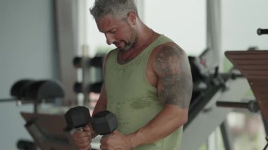 Bir adam spor salonunda dambıllarla antrenman yapıyor. Egzersiz sırasında odaklanmış ve kararlı görünüyor..