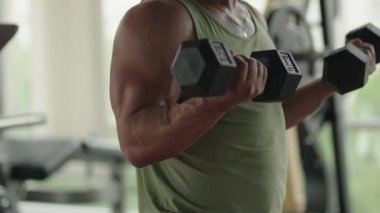 Yeşil kolsuz tişörtlü bir adam spor salonunda dambıllarla çalışıyor. Fitness, dayanıklılık eğitimi ve egzersiz kavramları için mükemmel..