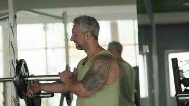 Dövmeli bir adam spor salonundaki halterden ağırlık alır. Forma, güce ve egzersiz rutinine odaklanıyor.
