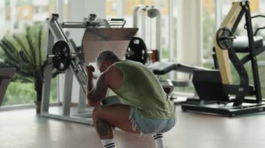 Yeşil kolsuz bluzlu ve kot şortlu bir adam modern ve iyi donanımlı bir spor salonunda çömelme egzersizi yapıyor..