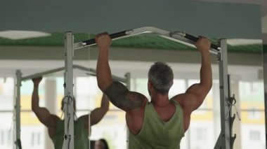Spor salonunda barfiks çeken bir adam. Güç antrenmanı ve fitness gösterisi yapıyor. Yeşil bir atlet giyiyor ve kolunda bir dövme var..