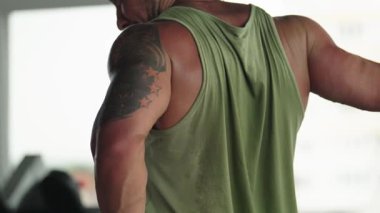 Dövmeli kaslı bir adam antrenmandan sonra kolunu uzatıyor. Yeşil bir atlet giyiyor ve odaklanmış görünüyor..