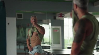 Orta yaşlı bir adam spor salonunun aynasında fiziğini gururla gözlemler. Antrenmanlarının sonuçlarını gösterir..