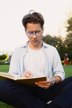 Odaklanmış genç bir adam çimenlerde oturuyor, kitap okuyor ve dışarıda notlar yazıyor..