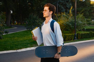 Gün batımında parkta elinde kaykay ve kitaplarla dolaşan genç bir adam, sıradan ve çalışkan bir hava yayıyor..