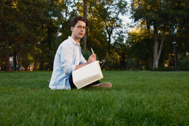 Çimlerin üzerinde oturan genç bir adam, yeşilliklerle çevrili, bir deftere yazıyor, düşünceli görünüyor..