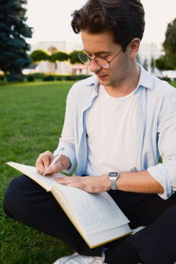 Parkta güneşli bir günün tadını çıkarırken açık bir günlüğe yazan yeşil çimlerin üzerinde oturan genç bir adam. Gelişigüzel ve rahat ayar odaklanma, yaratıcılık ve kişisel yansıma.