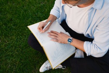 Çimlerin üzerinde oturan bir kişi dışarıda kalemle yazarken bir defter tutar. Ortam, sakin bir doğal ortamın altında yansıtıcı ya da yaratıcı bir an olduğunu gösteriyor..