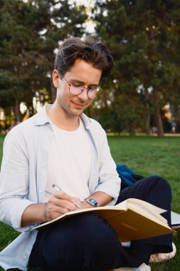 Genç bir adam çimenli bir alanda oturur, odaklanmış bir şekilde bir deftere yazar. Doğal çevreyle birlikte huzurlu bir ortamda yaratıcılık ya da çalışma anı yakalamak..