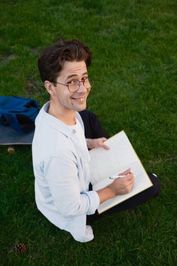 Çimenli bir tarlada oturmuş gülümseyen ve kameraya bakan neşeli genç bir adam.