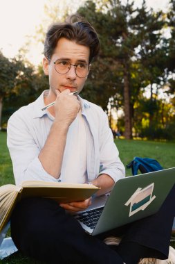 Odaklanmış genç bir adam çimenlerde oturuyor, bir dizüstü bilgisayar ve açık havada çalışıyor, düşüncelere dalmış, güneşli bir parkın doğal ortamının tadını çıkarıyor, eğitimi ve konsantrasyonu temsil ediyor.