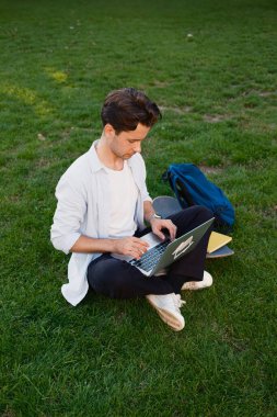 Odaklanmış genç bir adam, sırt çantası ve kaykayıyla çimenli bir açık alanda otururken dizüstü bilgisayarıyla modern yaşam tarzını ve açık hava verimliliğini gözler önüne seriyor..