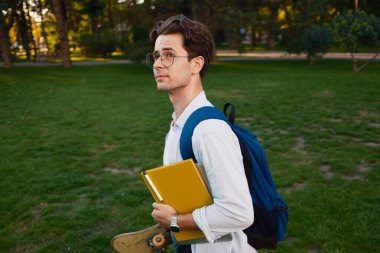 Sırt çantası takan genç bir erkek öğrenci parkın arka planına karşı kitap ve kaykay tutuyor..