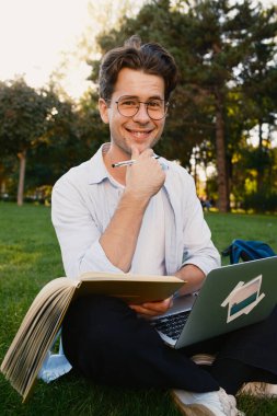 Gülümseyen genç adam parkta çimenlerde oturup dizüstü bilgisayar ve kitap okuyarak dingin ve entelektüel bir atmosfer yaratıyor.