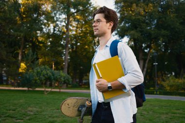 Yeşil bir parkta elinde kaykay ve not defteri olan genç bir adam boş zamanı ve eğitimi simgeliyor. Huzurlu ortam, keşif ve aktif yaşam tarzına katkıda bulunur.