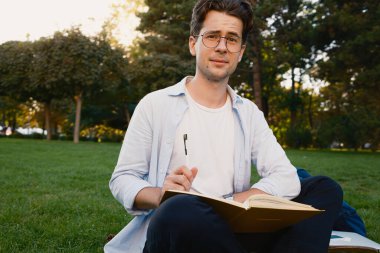 Parkta çimenlerde oturan, elinde açık bir defter tutan ve ders çalışan biri. Çevre yeşilliklerle doludur, barışçıl ve odaklanmış bir açık hava öğrenme ortamı göstermektedir..