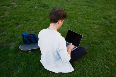 Genç bir adam yakınlarda kaykay ve çanta ile bir dizüstü bilgisayar kullanarak çimlerin üzerinde oturuyor. Açık havada..