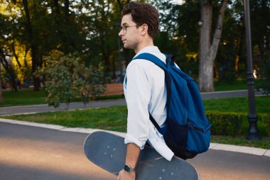 Gözlüklü ve sırt çantalı genç bir adam parkta yürürken bir kaykay tutuyor. Sahne, sakin ve cezbedici yeşil bir mekanın ortasındaki boş zamanı ve eğlenceyi resmediyor..