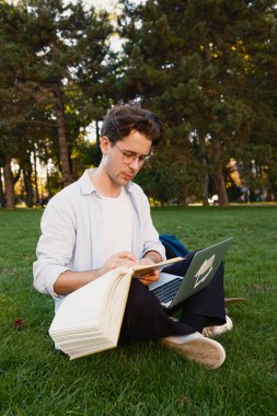 Genç adam parkta çimenli bir çimenlikte kitap ve dizüstü bilgisayarla çalışıyor, açık havada üretkenlik ve eğitim sunan, parlak bir gökyüzü altında yemyeşil ile çevrili..