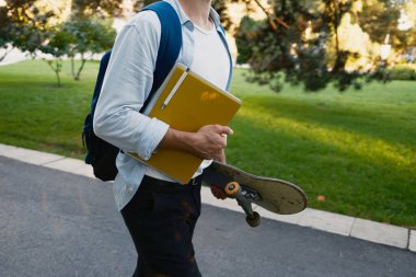 Sıradan giyimli bir öğrenci elinde kaykay ve yeşil defterle parkta yürüyor..